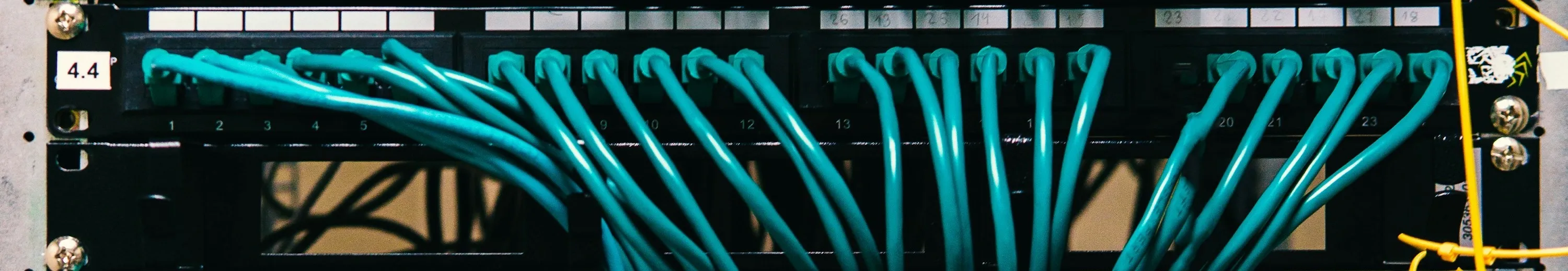 A patch panel with teal network cables in every port, its rows labelled by hand on strips of masking tape.