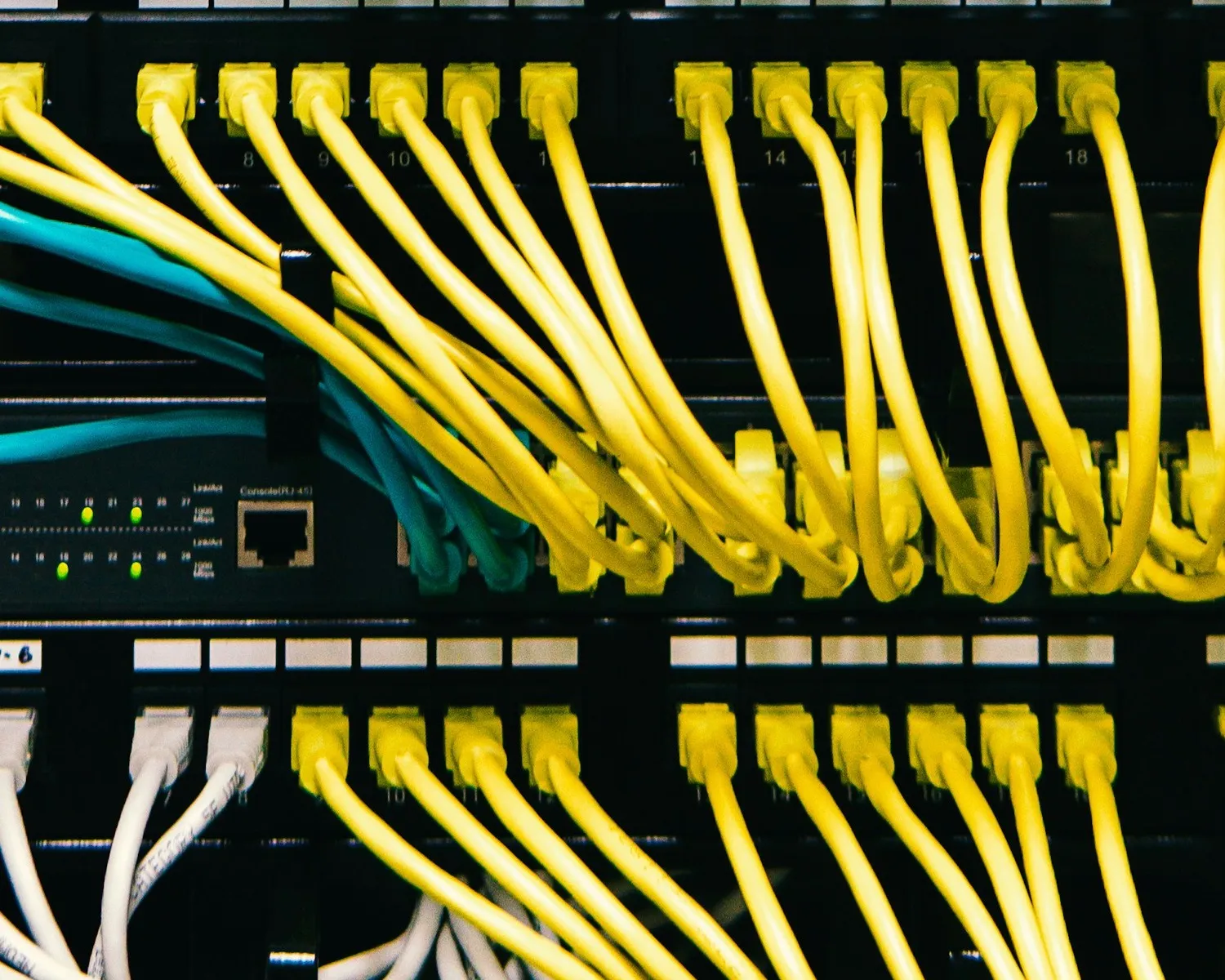 Yellow, teal, and white network patch leads converging into the numbered ports of a switch and patch panel.
